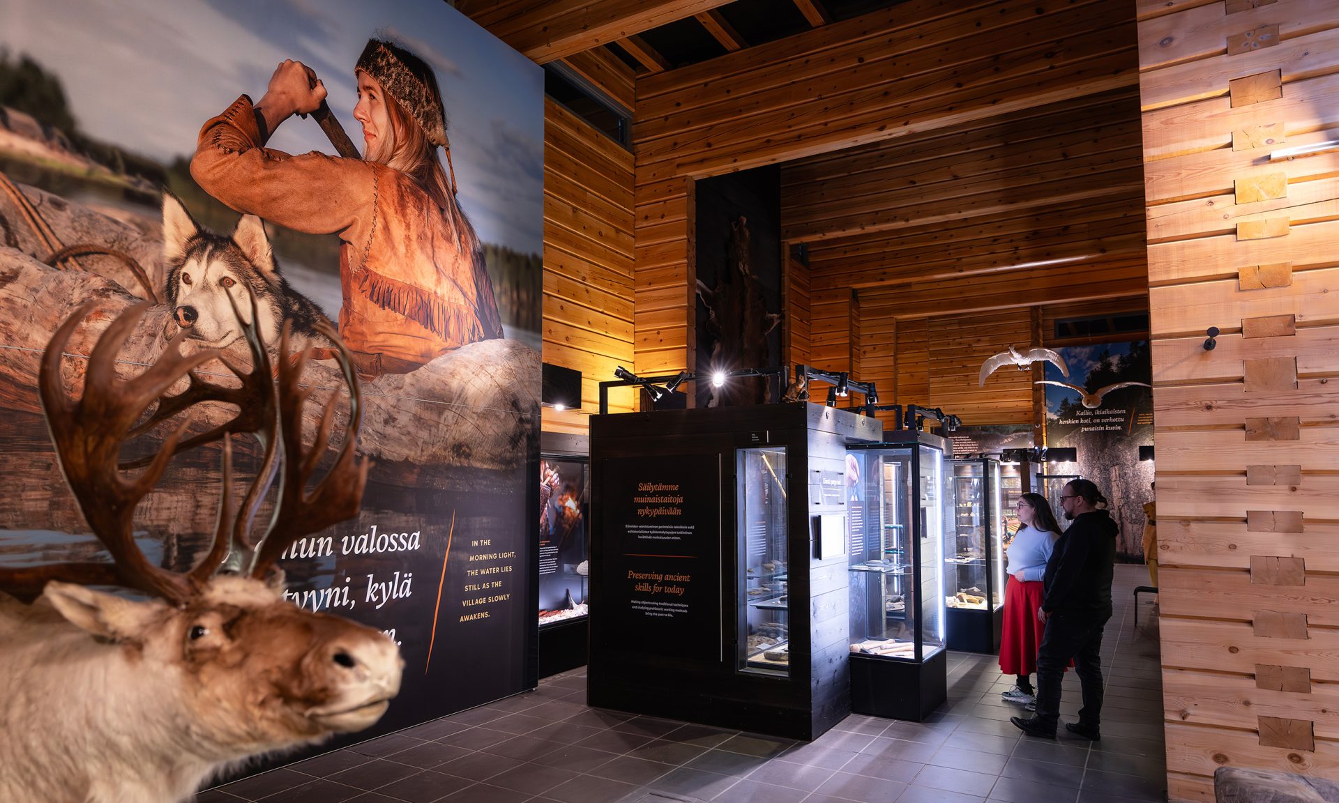 Kaksi henkilöä tutustumassa näyttelyvitriineihin. Etualalla on iso seinäkangas, jossa on kuva kivikautiseen asuun pukeutuneesta melojasta koirineen.