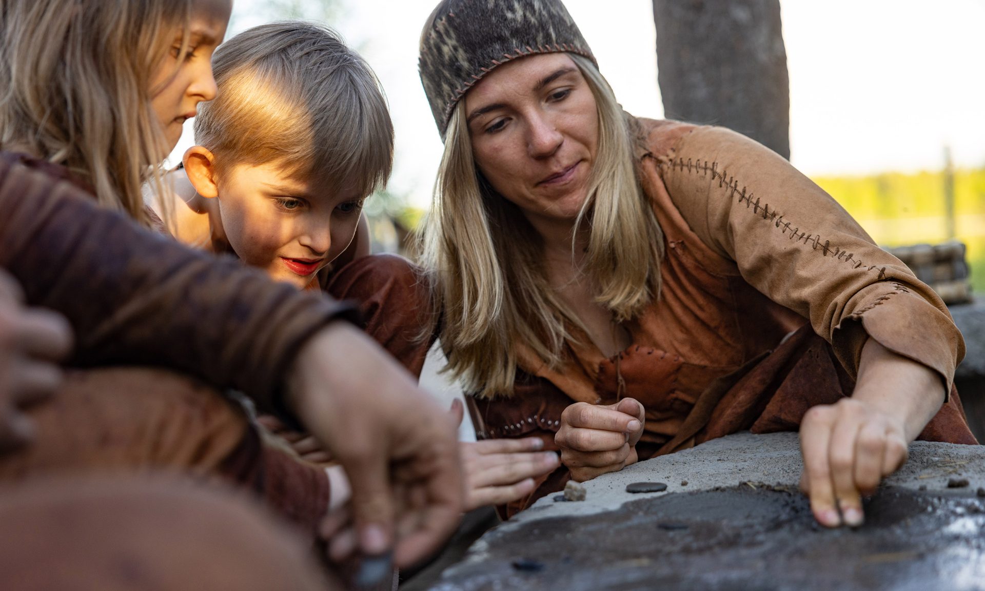 An adult is guiding two children in grinding a small stone into a piece of jewelry. All are dressed in Stone Age clothing.