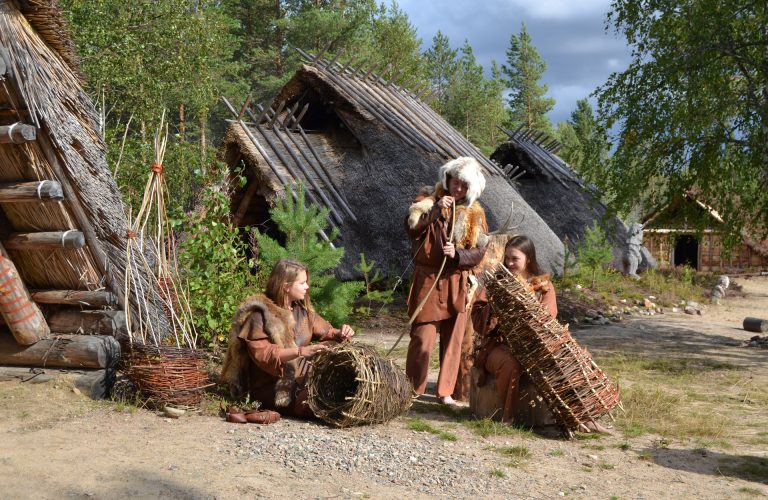 Nahkapukuihin pukeutuneita henkilöitä tekemässä pajutöitä. Takana näkyy kivikauden kylän asumuksia.