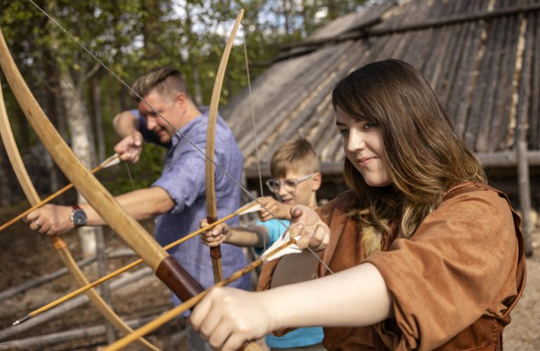 Three people shooting a bow. One of the people is wearing leather clothes. Stone Age Village can be seen in the background.