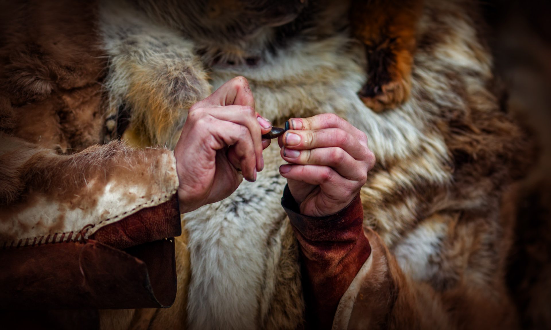 Close-up shot. A man dressed in Stone Age clothing grinds two small stones against each other.