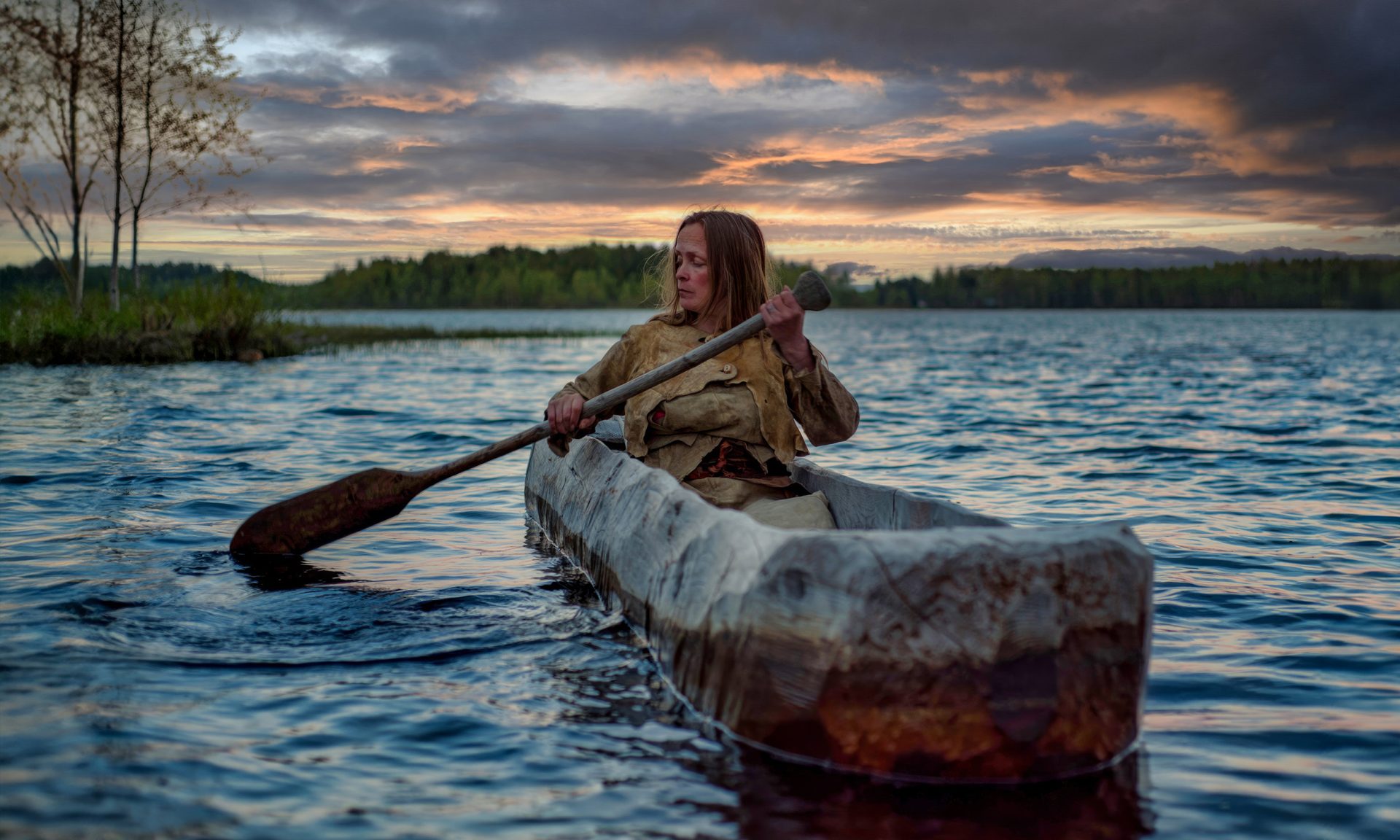 The guide, dressed in Stone Age accessories, paddles on a wooden, hand-carved rowboat. Summer evening, dim lighting.