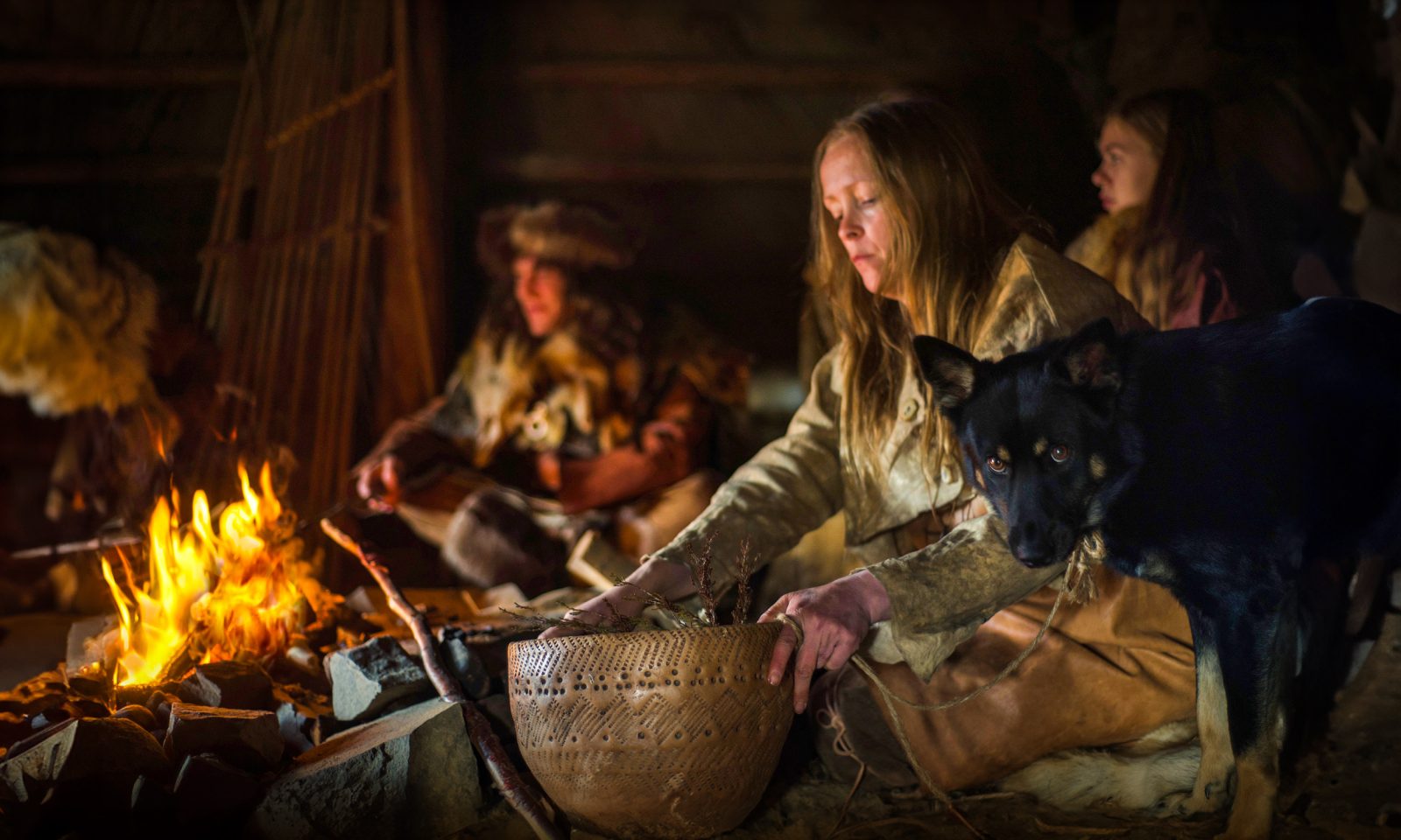 Interior view of a Stone Age dwelling. A woman cooks food in a large ceramic dish over a campfire.