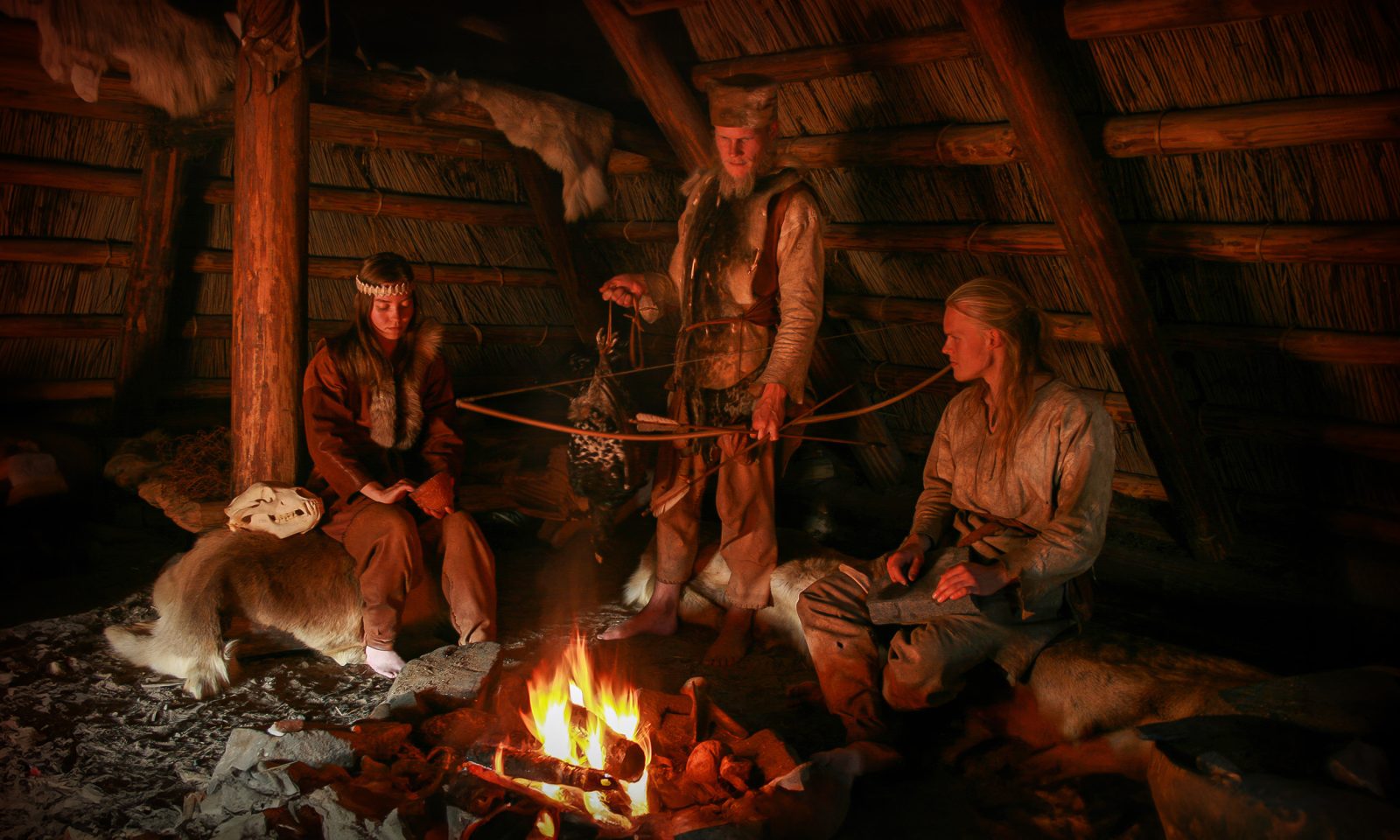 Guides in Stone Age clothing inside the restored dwelling by the campfire. Dim lighting.