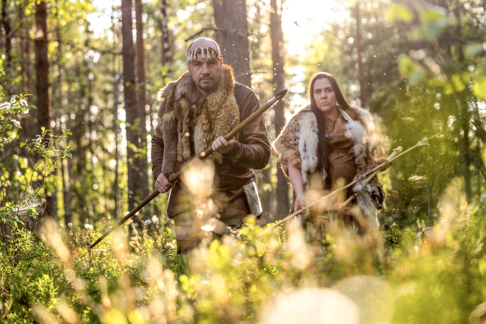 Kaksi kivikautisiin nahkavaatteisiin pukeutunutta henkilöä hiipivät metsässä aseet käsissään.