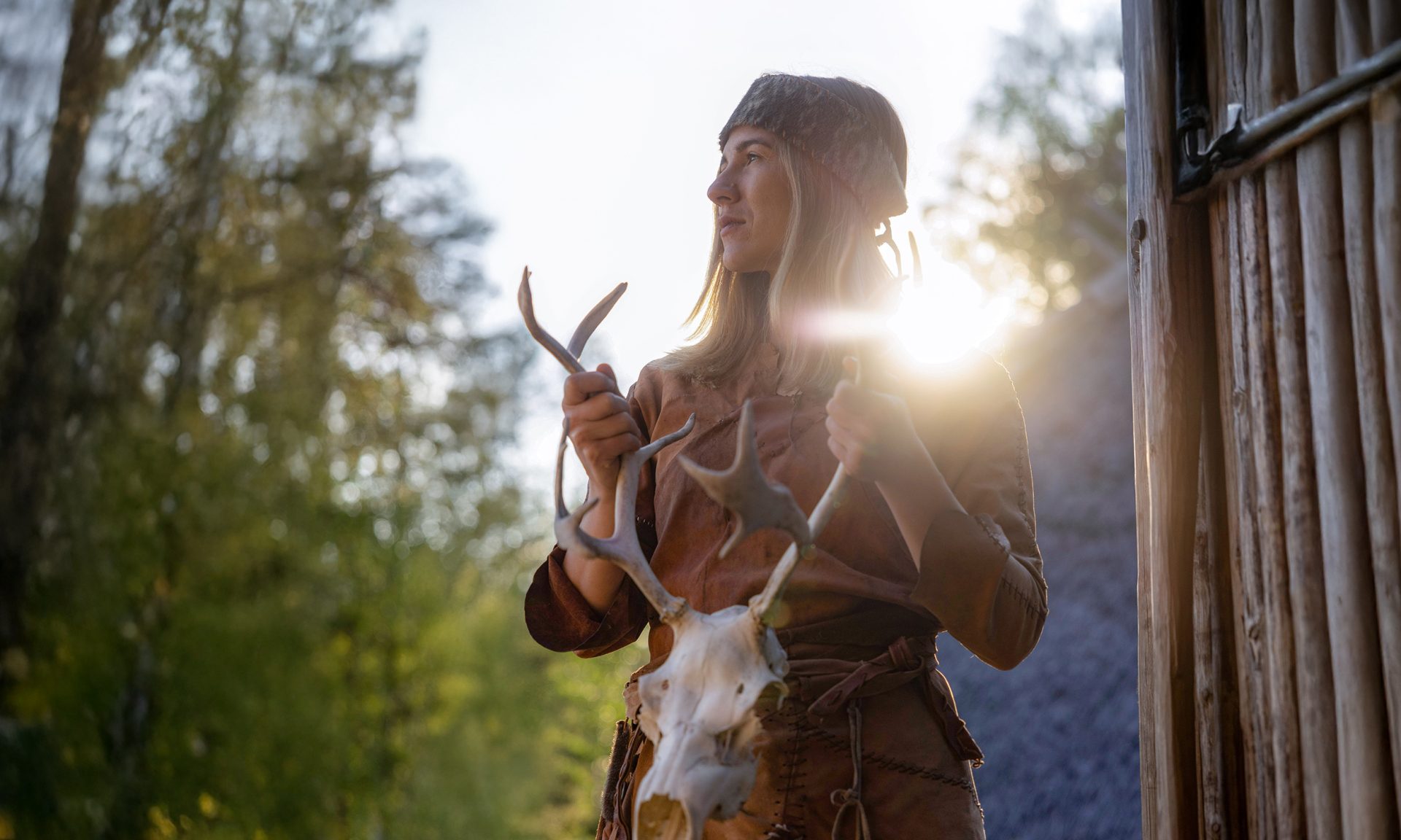 Kivikauden nahkavaatteisiin pukeutunut nainen pitää käsissään poron kalloa. Kesäilta, aurinko loistaa naisen takaa.