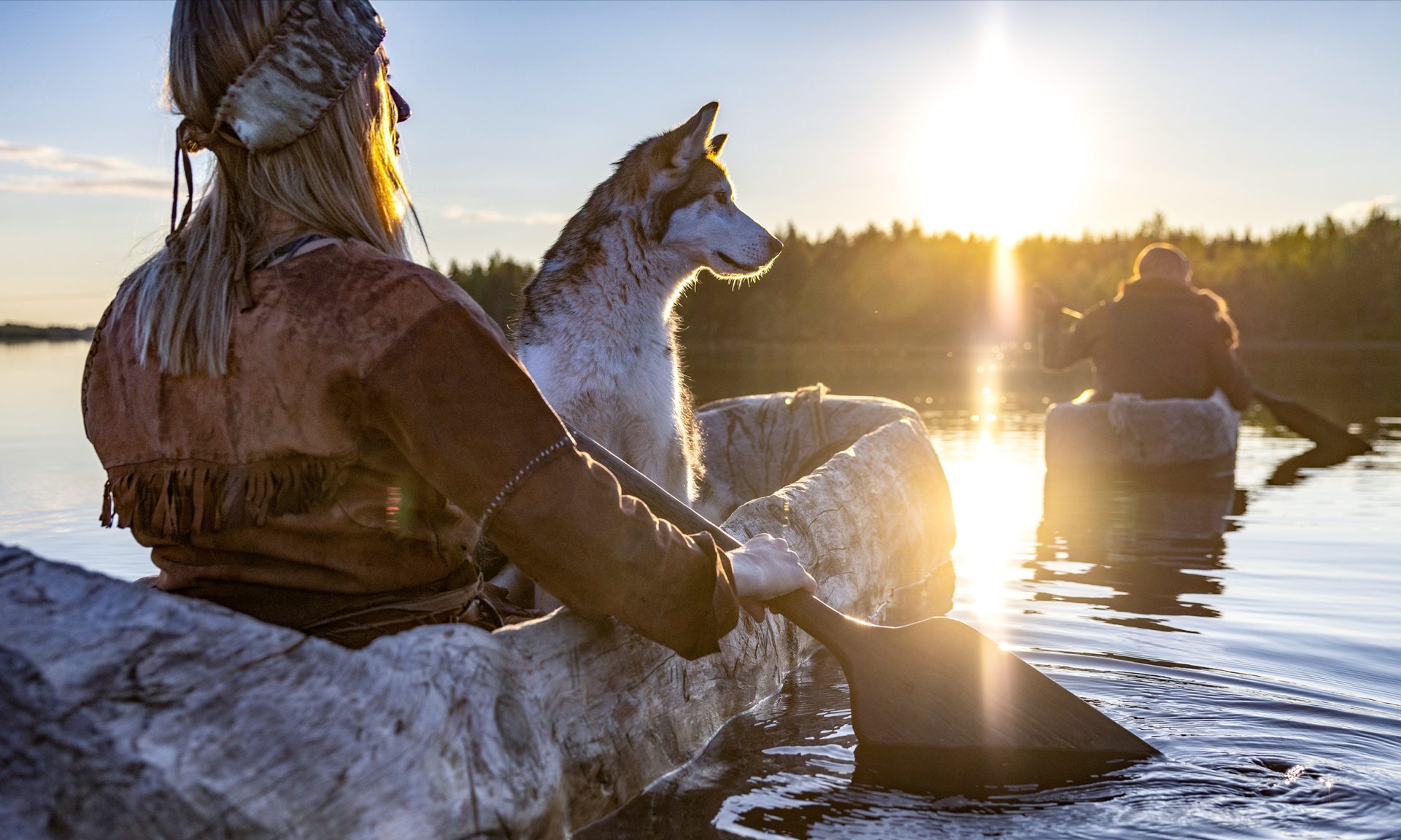 Kivikauden asuun pukeutunut nainen meloo puisella ruuhella järvellä koira kyydissään. Kesäilta, laskeva aurinko loistaa puiden takaa järven laidalla