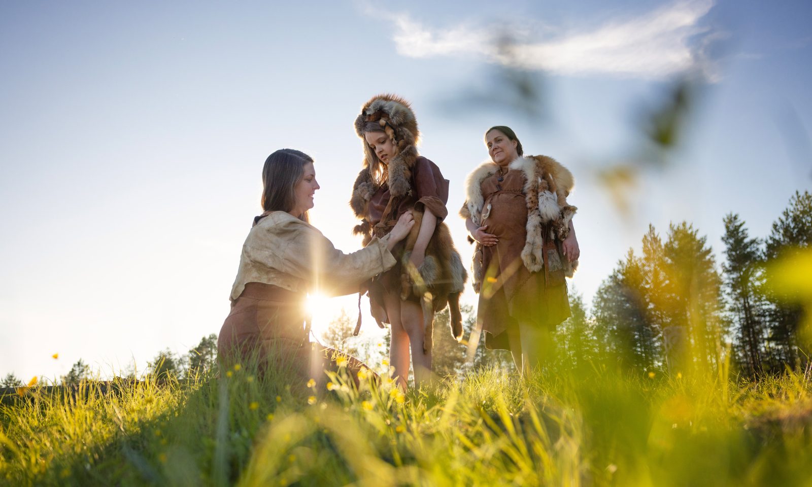 Kolme henkilöä kivikautisissa asuissa seisoo kesäisellä niityllä ilta-auringon valossa, etualalla nainen pukee turkiksia lapsen ylle.