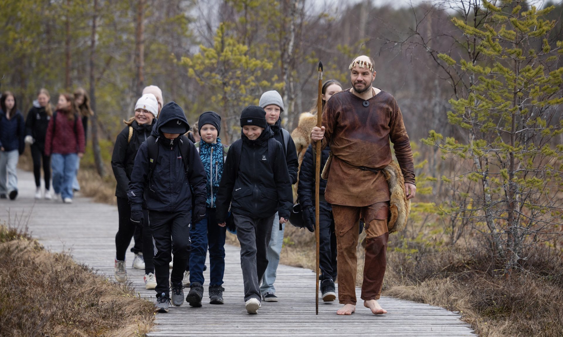 Kivikauden nahka-asuun pukeutunut henkilö johtaa koululapsia puisella kävelyreitillä metsässä. Miehellä on keihäs kädessään.