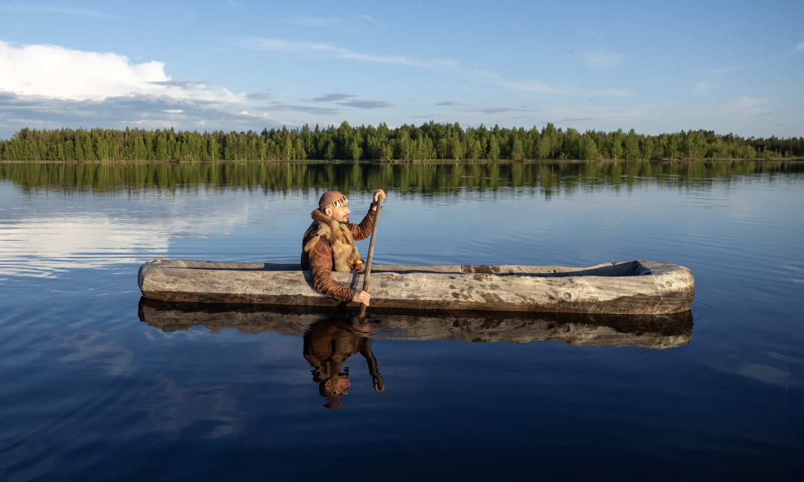 A person dressed in Stone Age clothing is paddling a wooden paddle. It's a summer day, the water is calm.