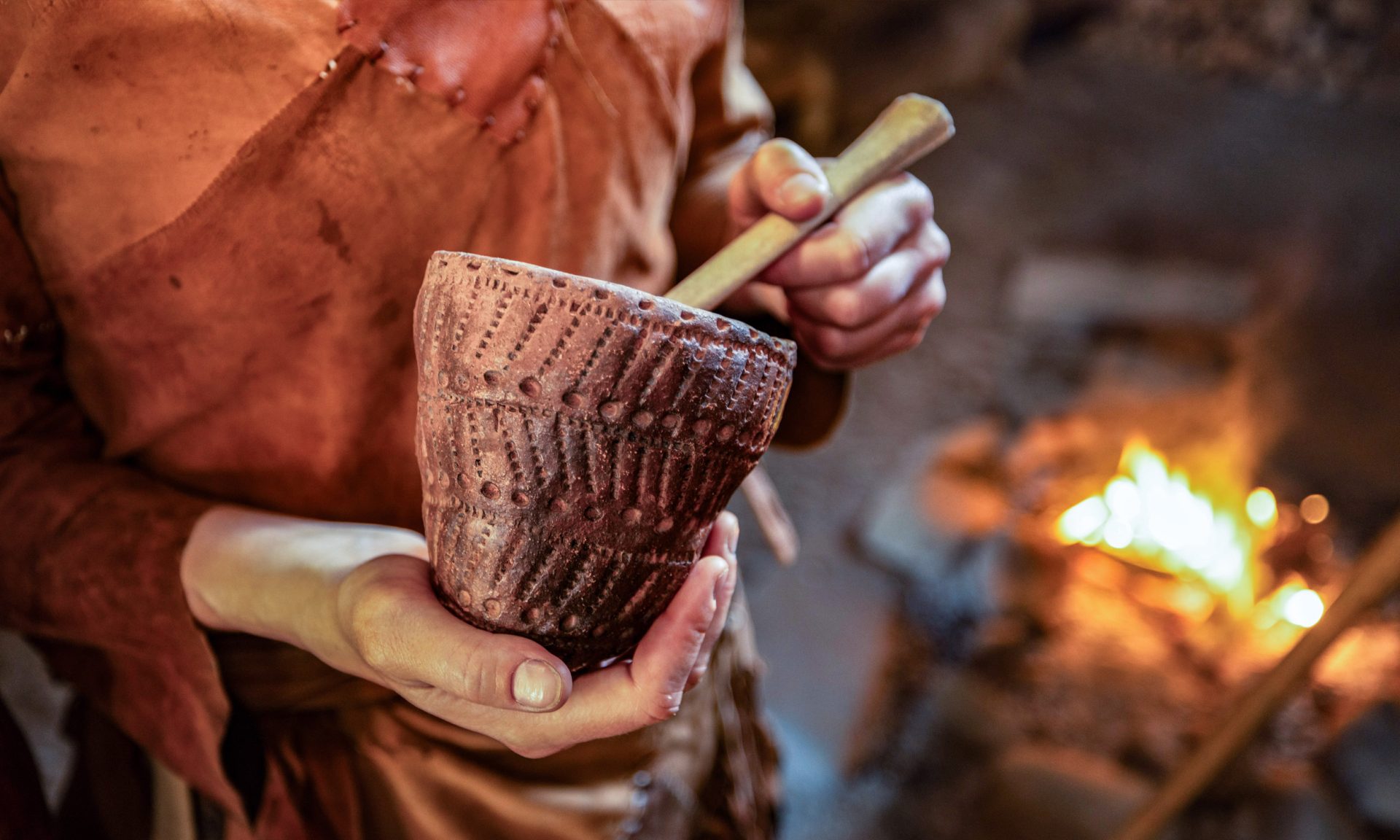 Close-up. A person dressed in Stone Age clothing holds a clay pot in his hand and stirs the contents with a ladle. A campfire can be seen in the background.