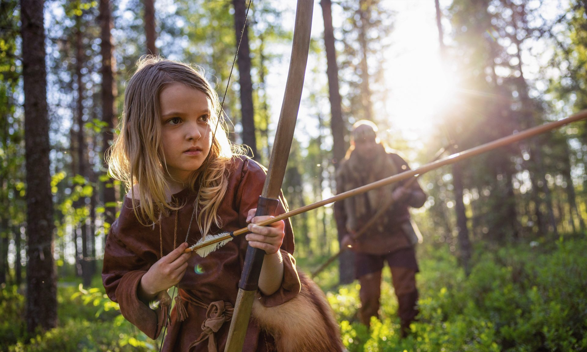 A child dressed in Stone Age clothing is stringing an arrow on a bow in a forest gilded by the evening sun, with a person holding a spear in the background.