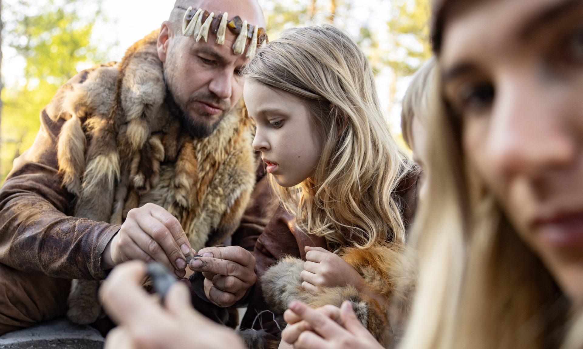 An adult instructs a child to polish a small stone into a piece of jewelry. Both are dressed in Stone Age clothing.