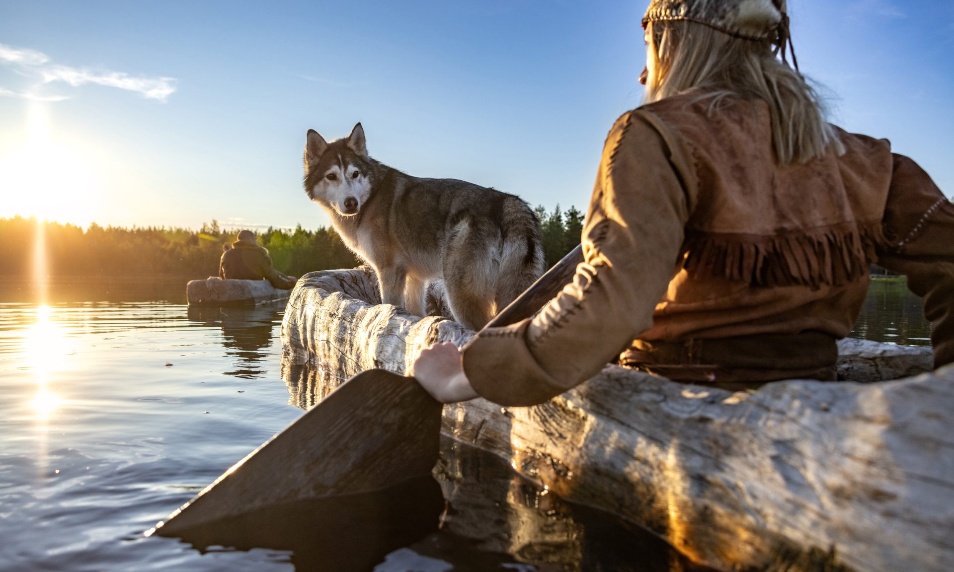 Kivikauden asuun pukeutunut henkilö meloo puisella ruuhella joella, jonka pinta on tyyni. Ruuhessa on myös koira, joka katsoo omistajaansa. Ilta-aurinko kultaa veden pinnan.