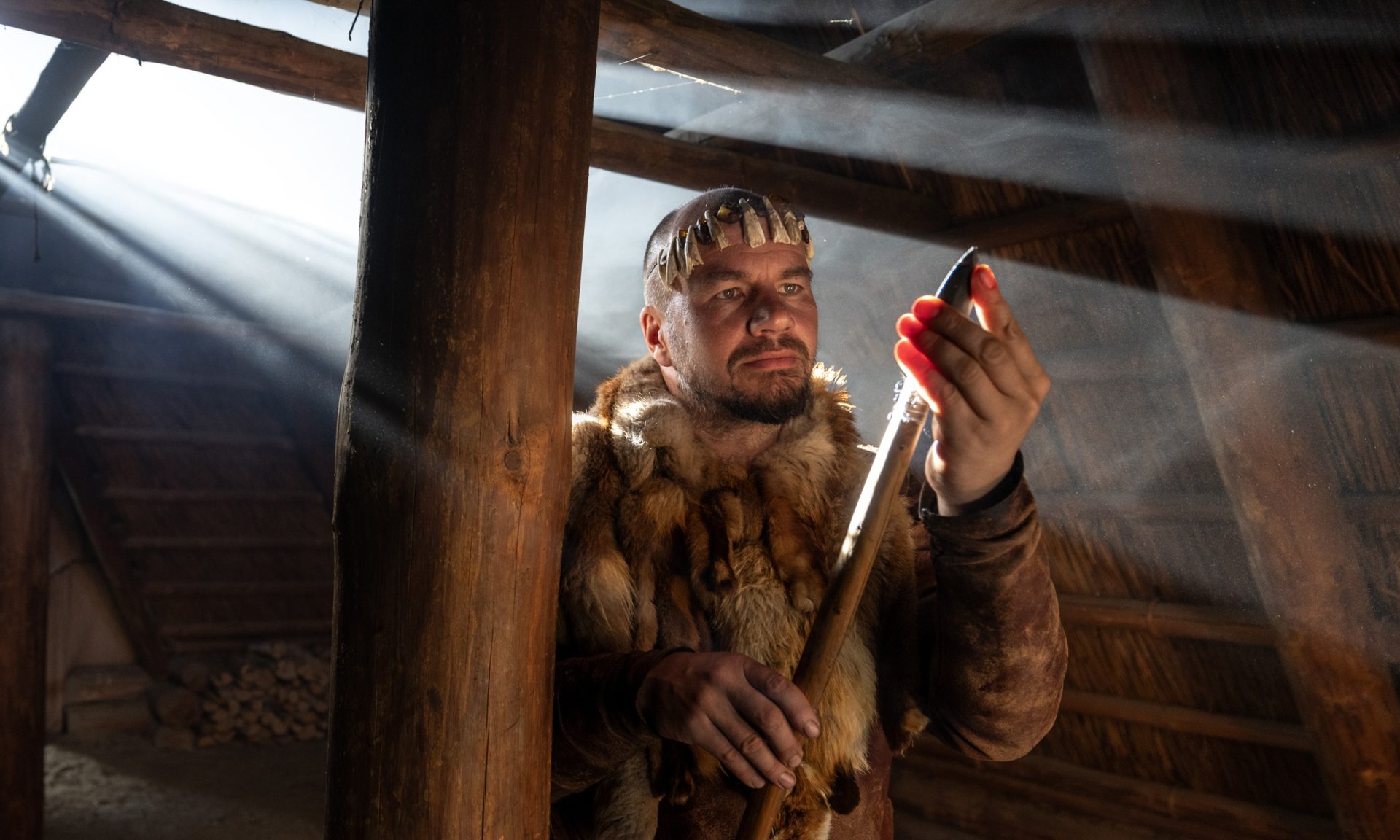 A person dressed in Stone Age clothing examines the sharpness of a spear's stone tip inside a Stone Age dwelling, where a beam of light shines beautifully.