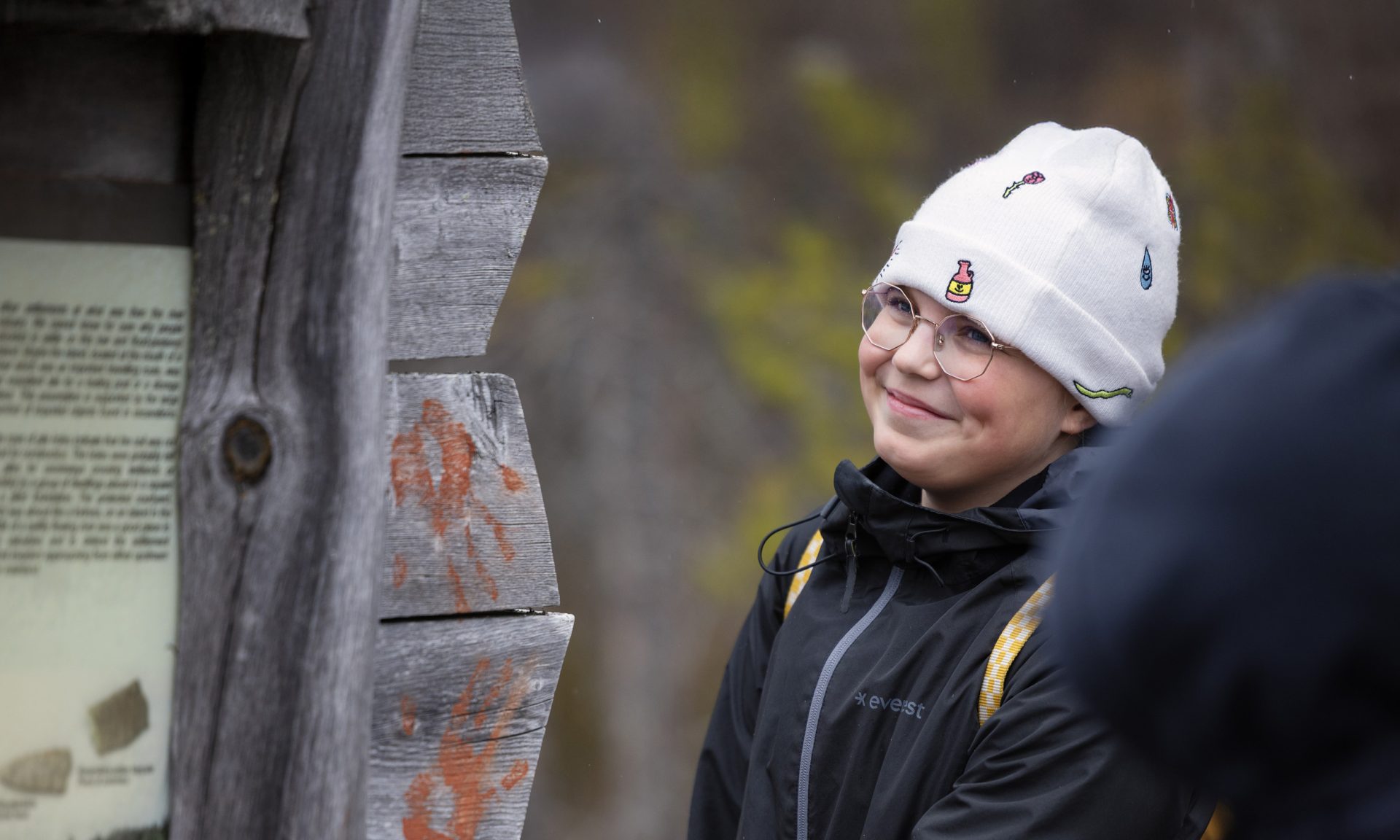 Lähikuva hymyilevästä lapsesta, joka seisoo puurakenteisen opaskyltin vieressä. Lapsella on yllään valkoinen pipo ja tumma ulkoilutakki.