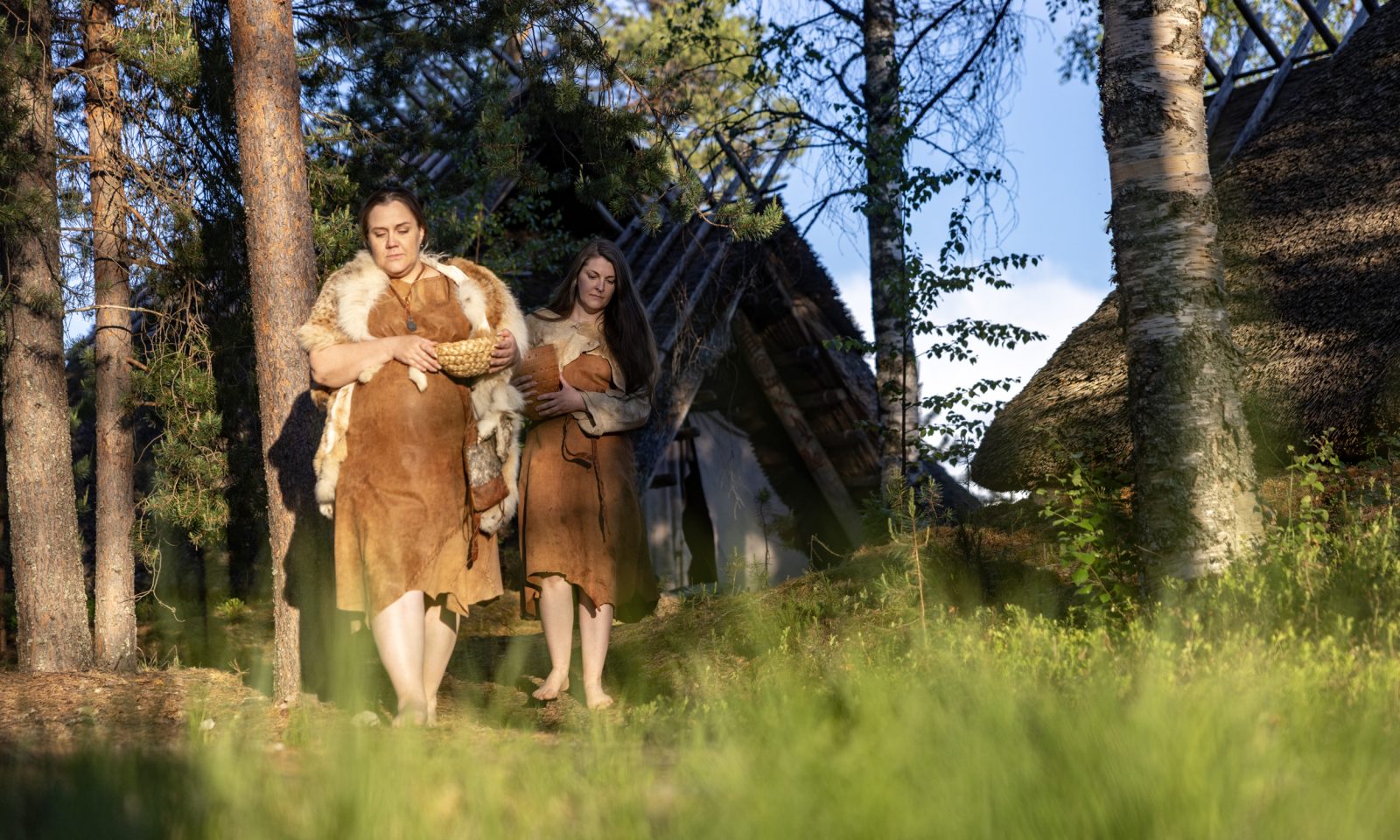 Two women dressed in Stone Age clothing are walking in the forest in the ancient village of Kierikki, with a Stone Age dwelling visible in the background.