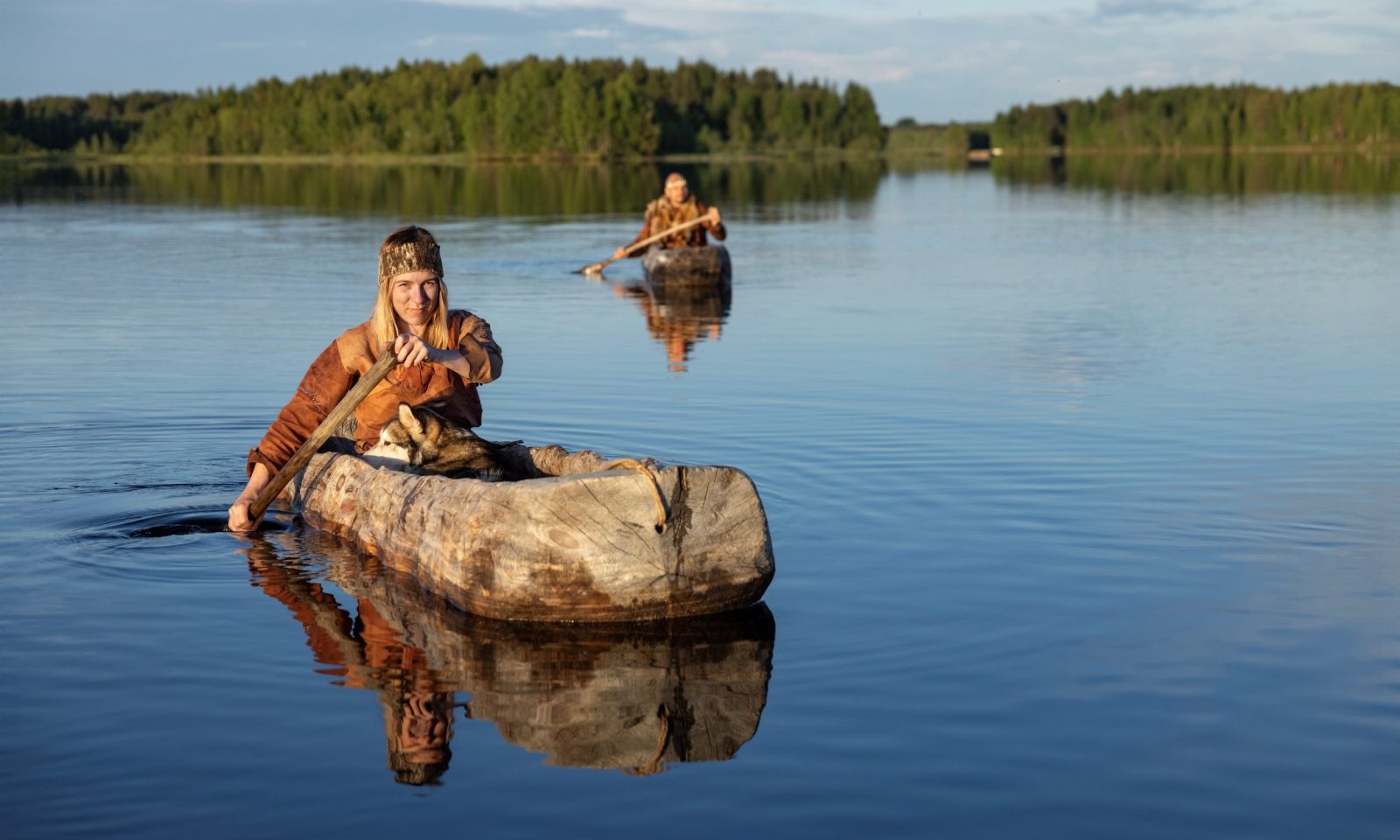 Nainen meloo puisella ruuhella, tyyni joki. Taustalla toinen meloja.