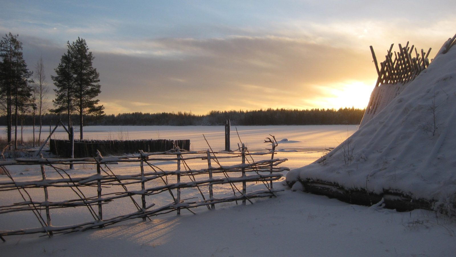 Talvi, etualalla vanha seipäistä rakennettu aita sekä kivikautisen rakennuksen nurkkaa. Aurinko paistaa, lunta on maassa.
