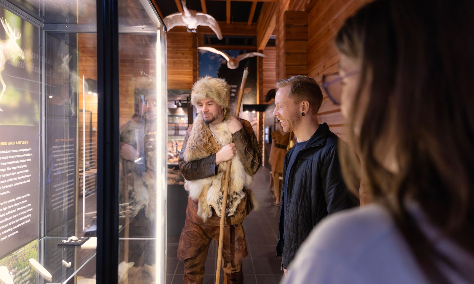 A presenter dressed in Stone Age clothing is telling two visitors about the contents of the display case. There are stuffed swans hanging from the ceiling of the exhibition space.